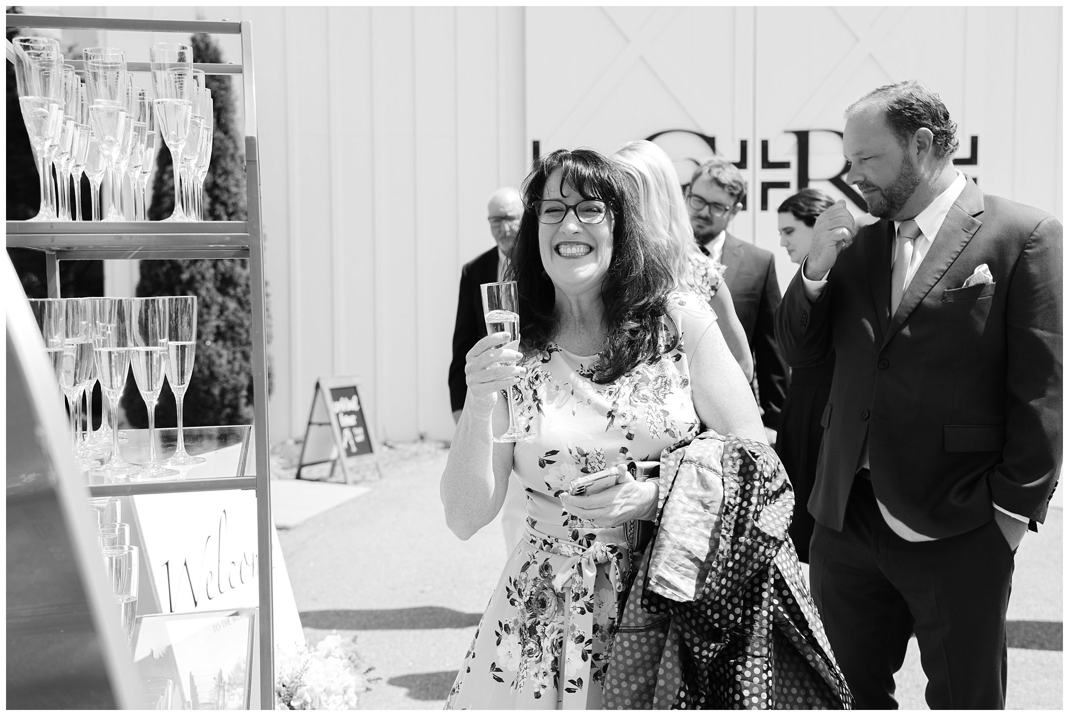 A woman in a floral dress stands smiling, holding a glass of champagne with a jacket draped over her arm. People in formal attire stand behind her near a shelf with more filled champagne glasses.