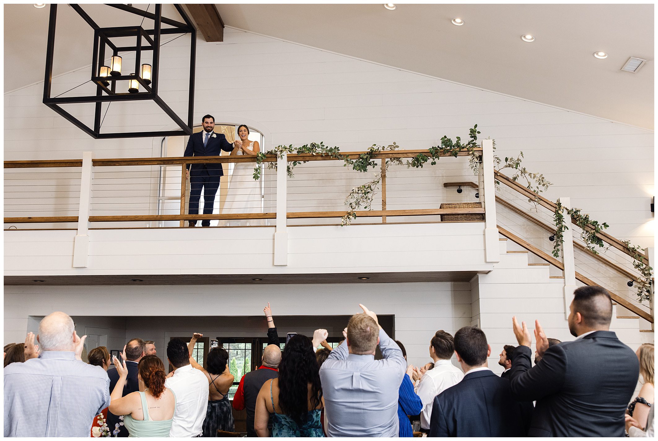 A couple stands on an elevated walkway, greeting a crowd of people below. The crowd is clapping and looking up at them. The walkway is decorated with greenery.