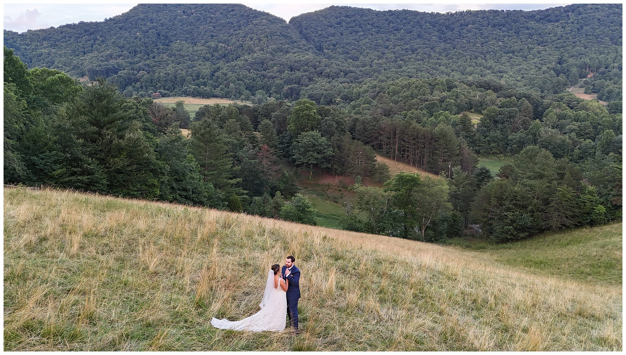 A bride and groom stand together on a grassy hilltop, surrounded by lush trees and mountains in the background.