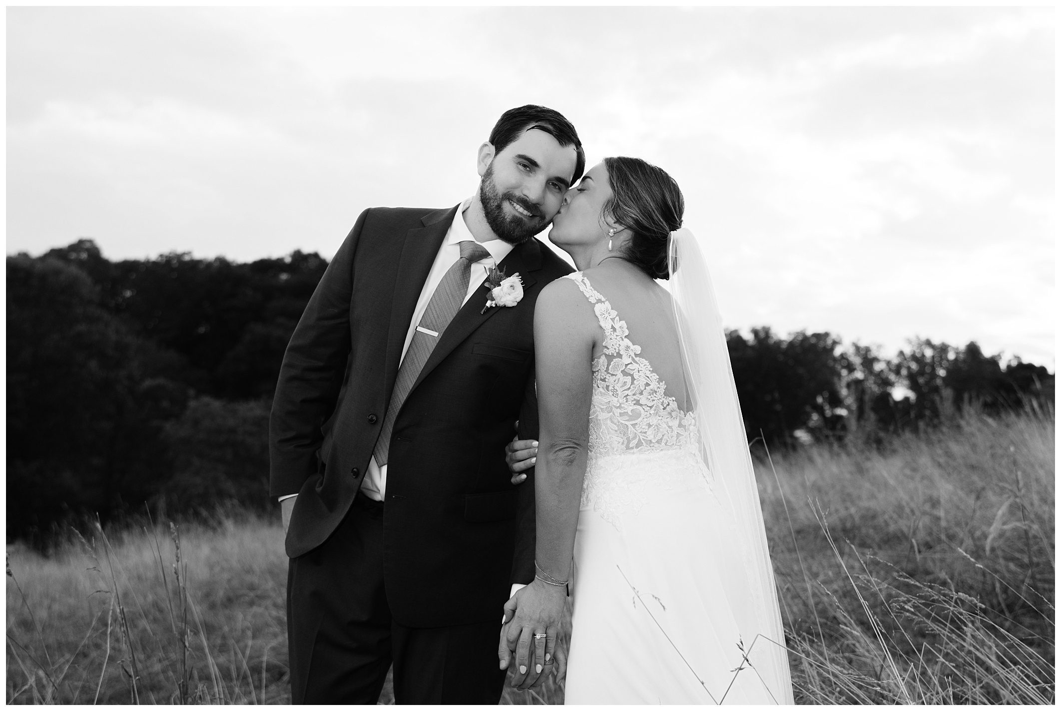 A bride and groom stand in a grassy field. The bride, in a white dress and veil, kisses the groom's cheek as they hold hands. The groom wears a suit with a flower boutonniere. Trees are visible in the background.