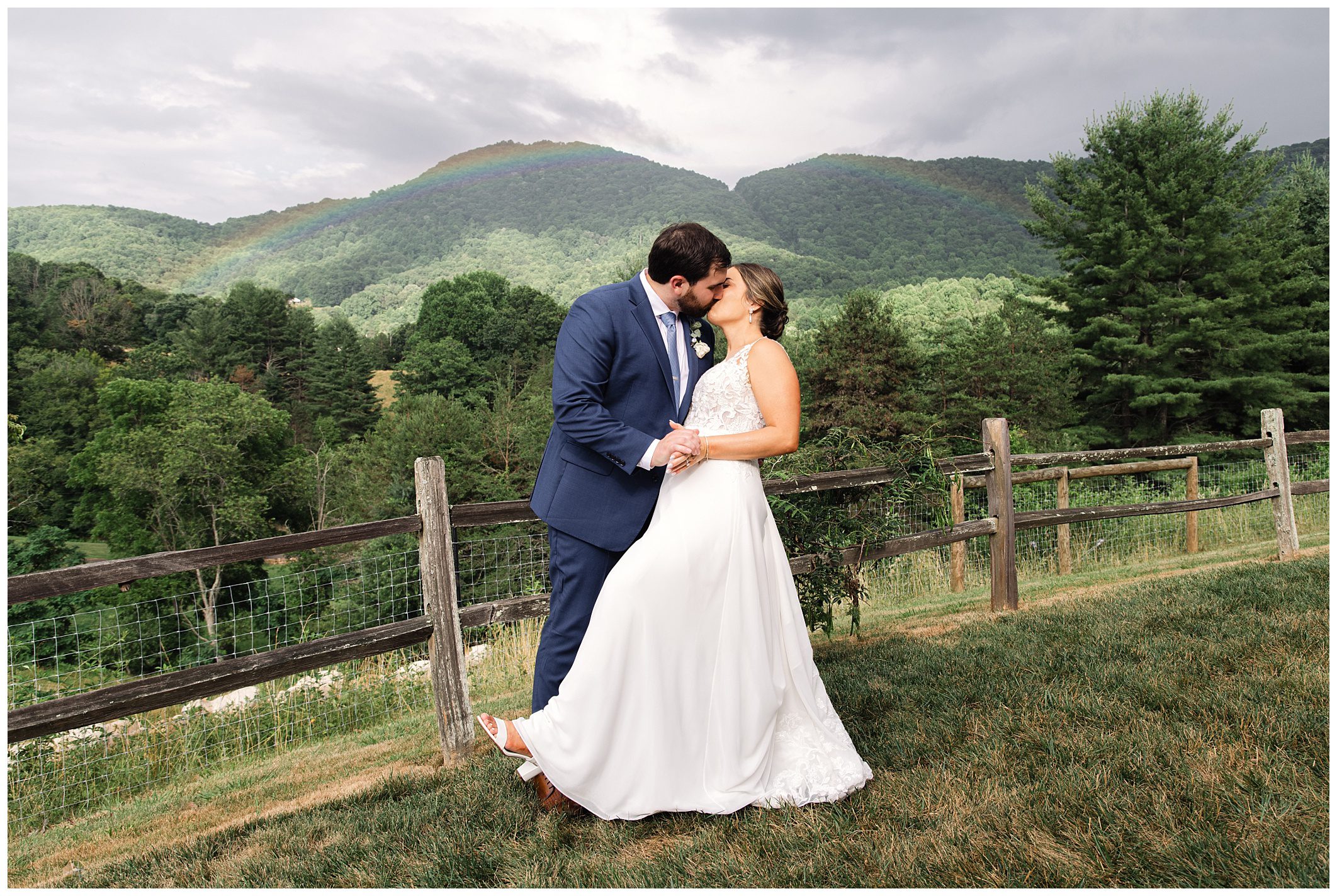 A couple in wedding attire kisses outdoors near a rustic fence with green hills and a faint rainbow in the background.