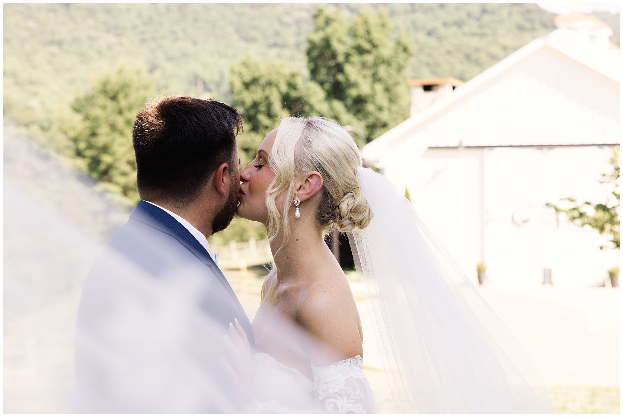 Elegant NC Wedding with A bride wearing a veil kisses a groom outdoors in front of a light-colored building with greenery in the background.