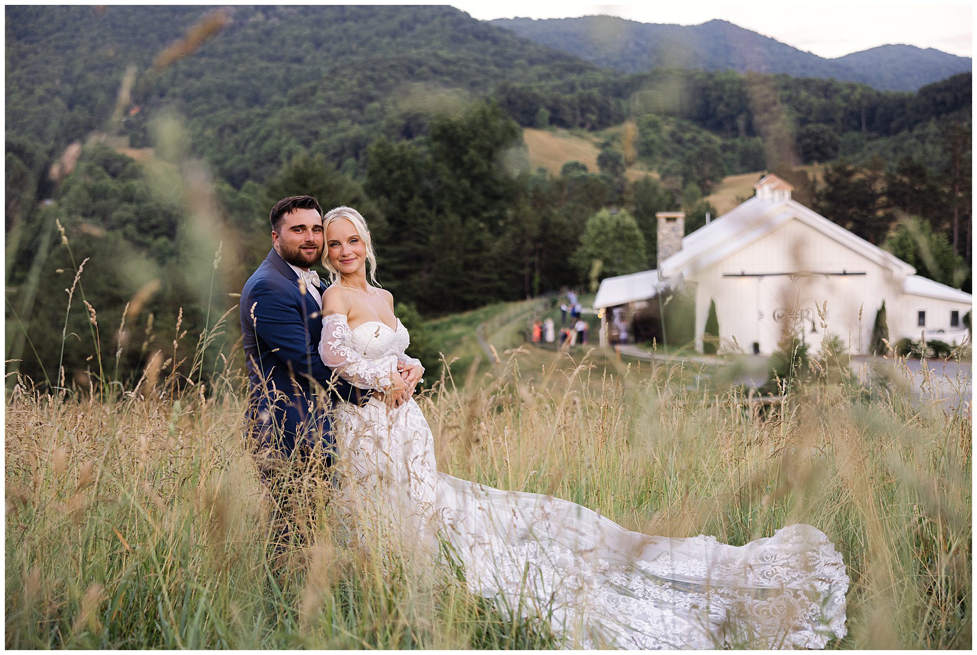 A bride and groom stand in a grassy field in front of a white building with long sleeves and outdoor ambiance, embracing and looking at the camera. Mountains and trees are visible in the background.