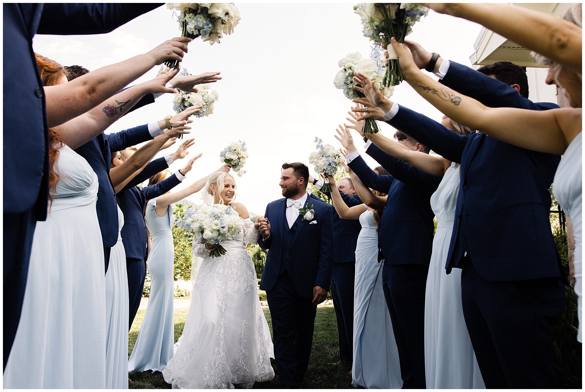 A bride and groom walk under an arch of raised bouquets held by bridesmaids and groomsmen dressed in coordinating outfits. The couple holds hands and smiles as they proceed.