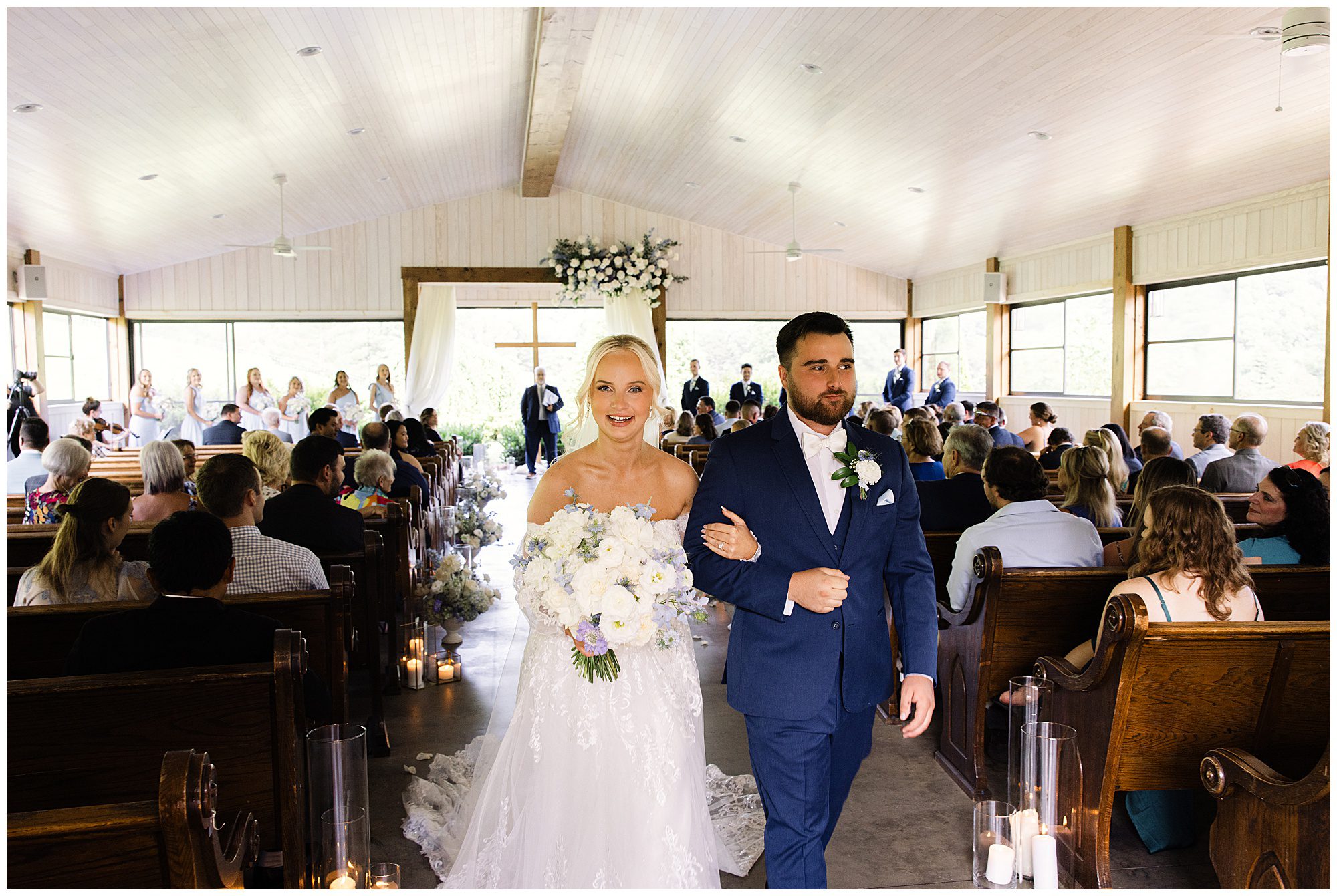 A newly married couple walks down the aisle, smiling and holding hands, in a wooden church filled with guests. The bride wears a white gown and holds a bouquet, while the groom is in a blue suit.