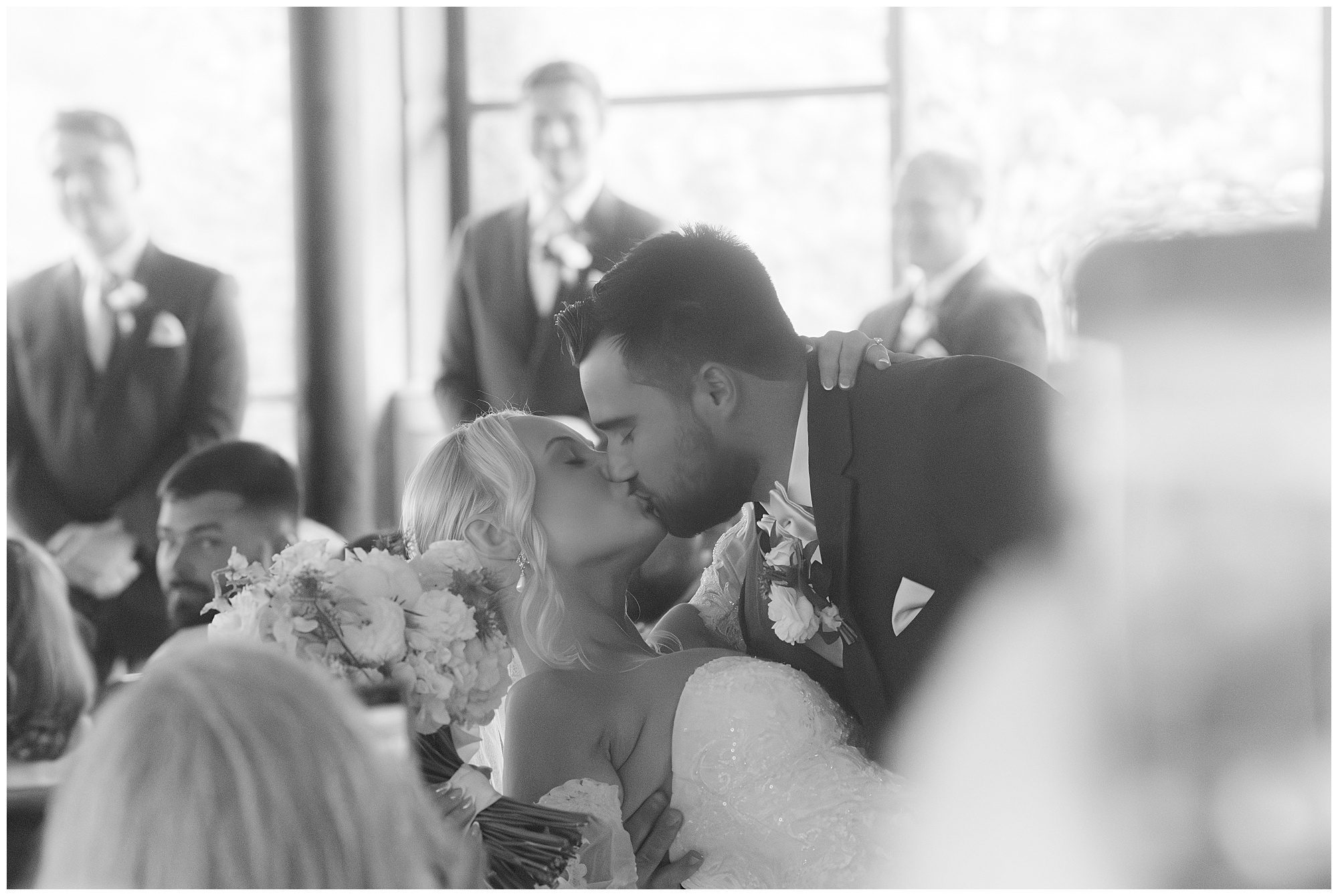 A bride and groom share a kiss at their wedding ceremony, surrounded by blurred guests and groomsmen in the background. The bride holds a bouquet of flowers. The setting appears soft and dreamlike.