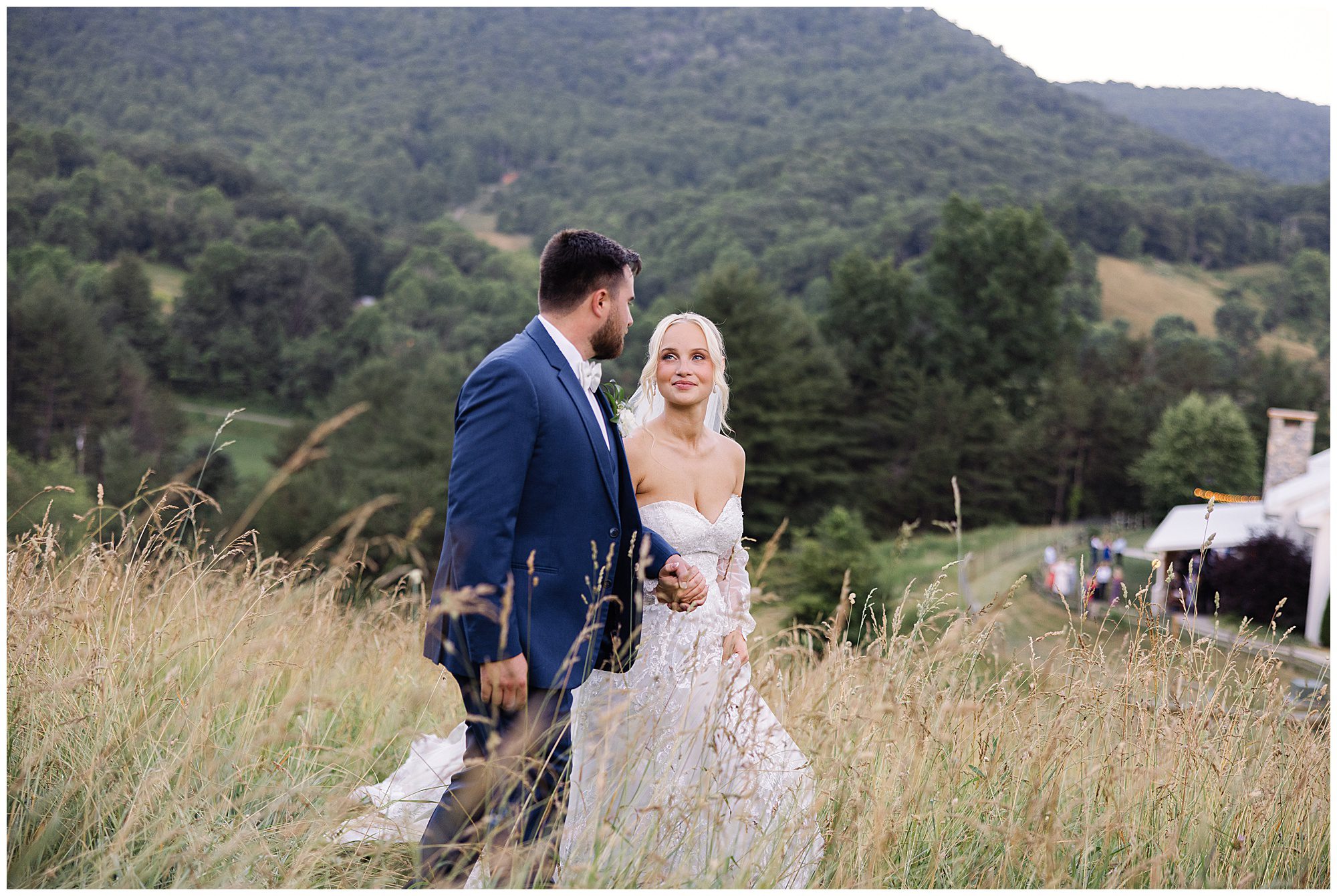 Bride and groom holding hands, walking through a grassy field with a forested mountain landscape in the background.