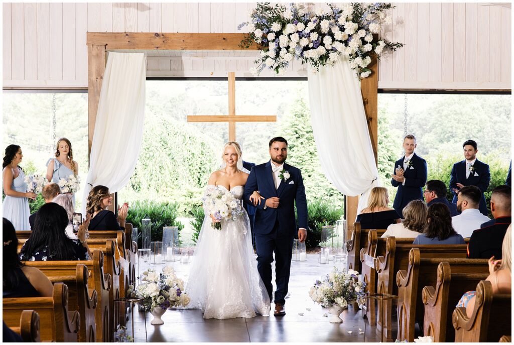 A newlywed couple walks down the aisle in a church decorated with white flowers and draped fabric. Guests are seated in pews, and a wooden cross is visible in the background.