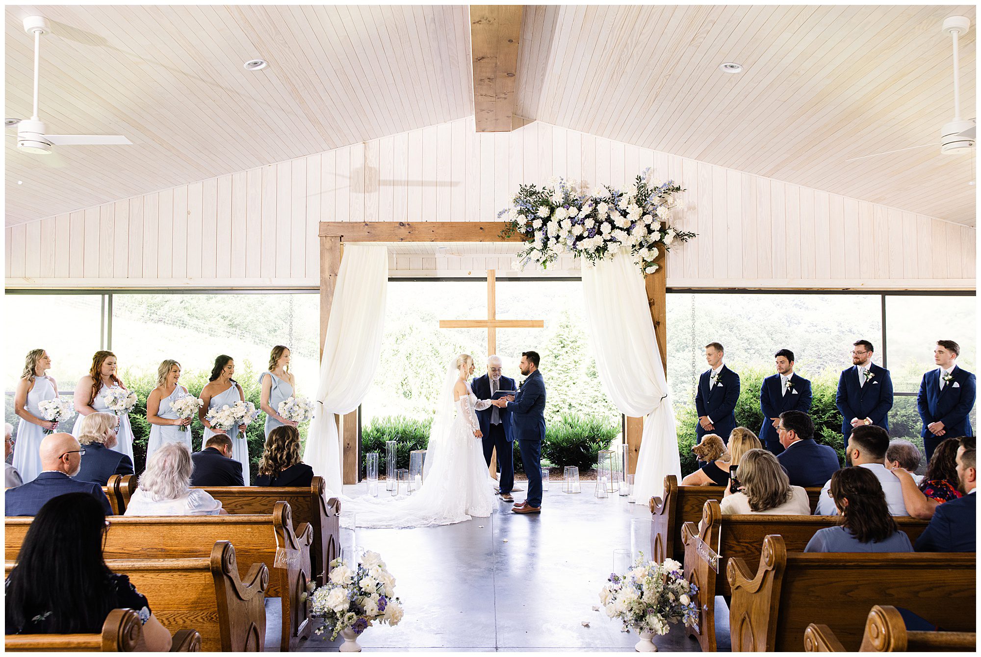 A couple stands at the altar exchanging vows in a bright, wood-accented chapel with a large cross, surrounded by bridesmaids in light blue dresses and groomsmen in navy suits, with guests seated in pews.