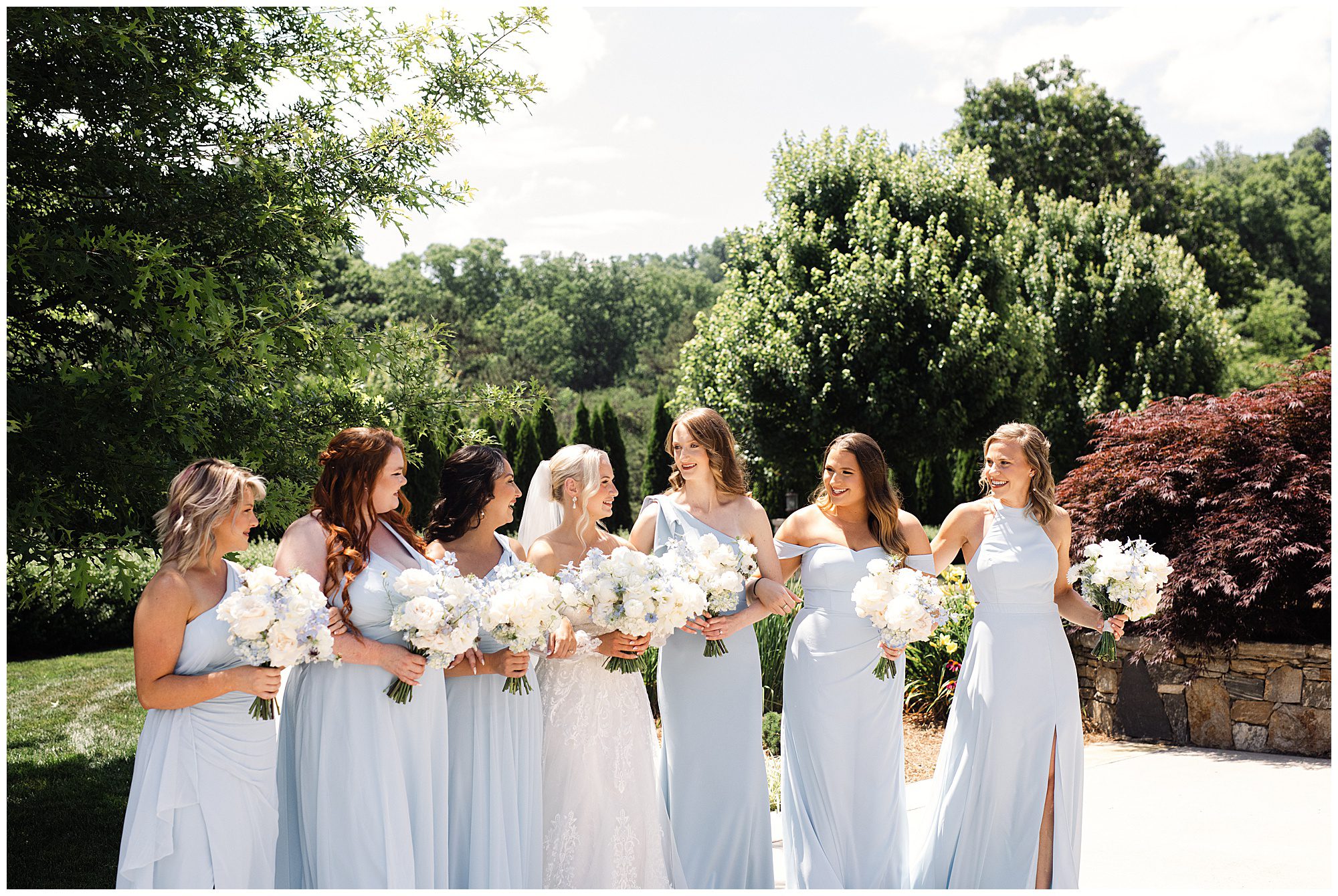 A bride in a white dress stands outdoors with six bridesmaids wearing light blue dresses, all holding white floral bouquets, against a backdrop of trees and shrubs.