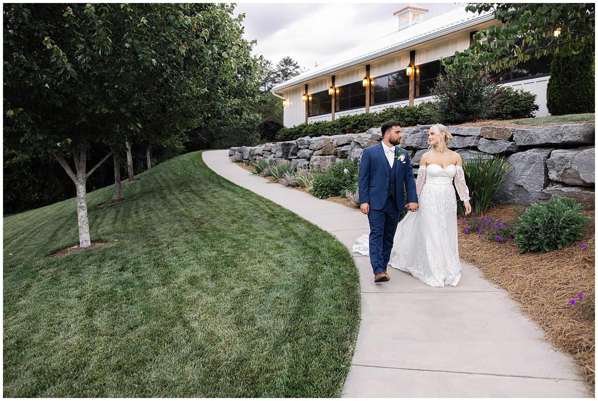 A bride and groom walk hand-in-hand on a curved path near a landscaped lawn and stone wall, with a well-lit building in the background.