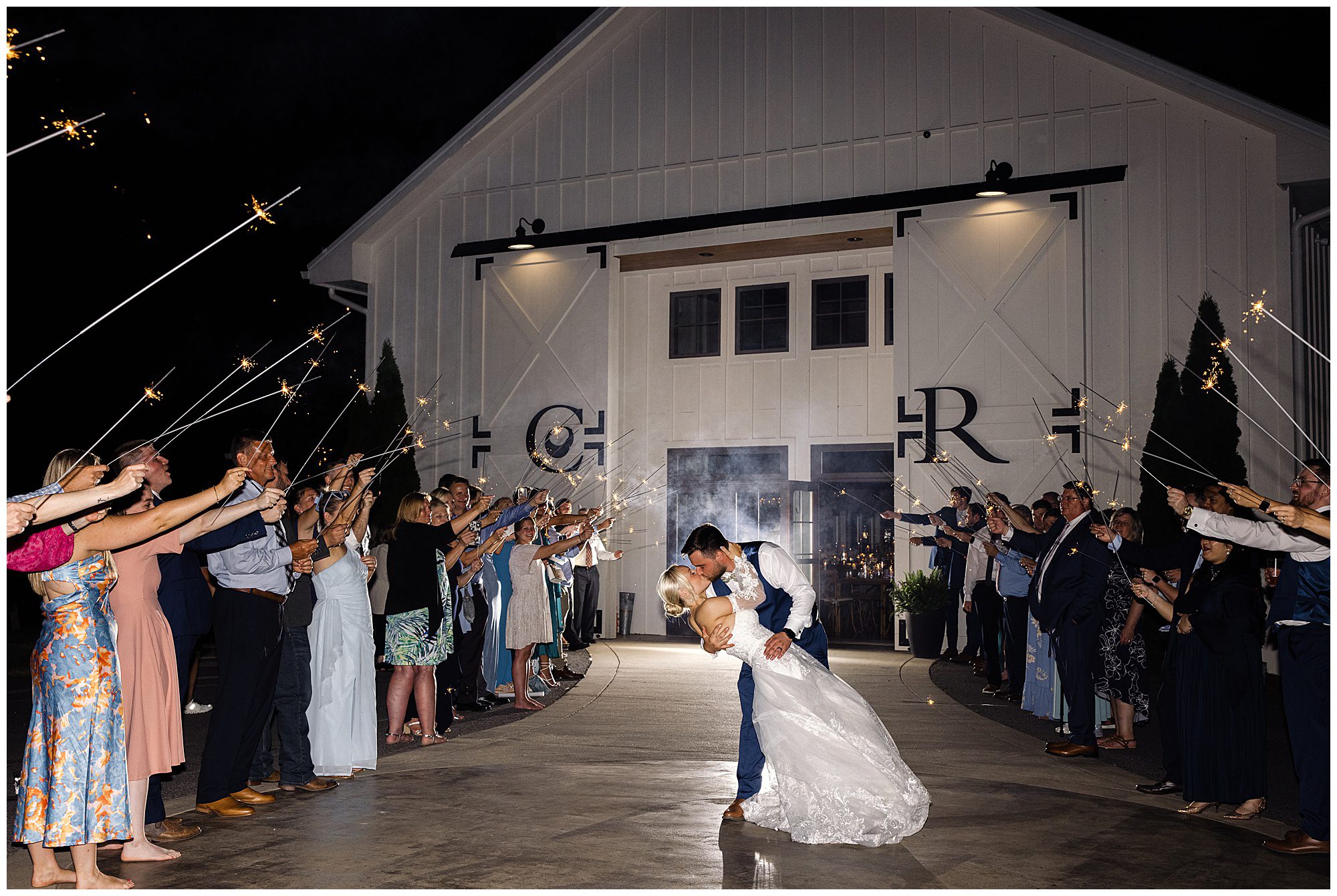 A couple embraces in a dramatic kiss surrounded by family and friends holding sparklers outside a white barn-style building at night.