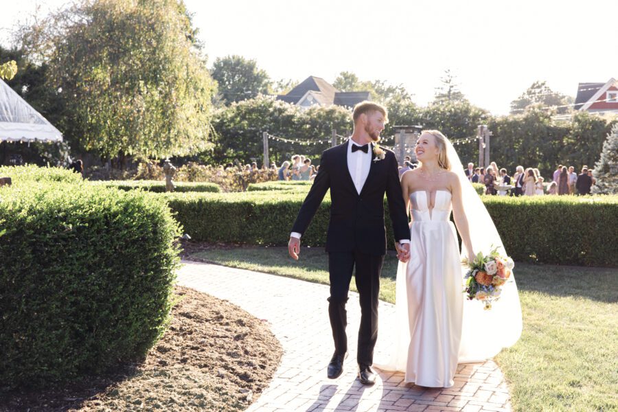 couple holding hands and walking together with guests at cocktail hour in the background