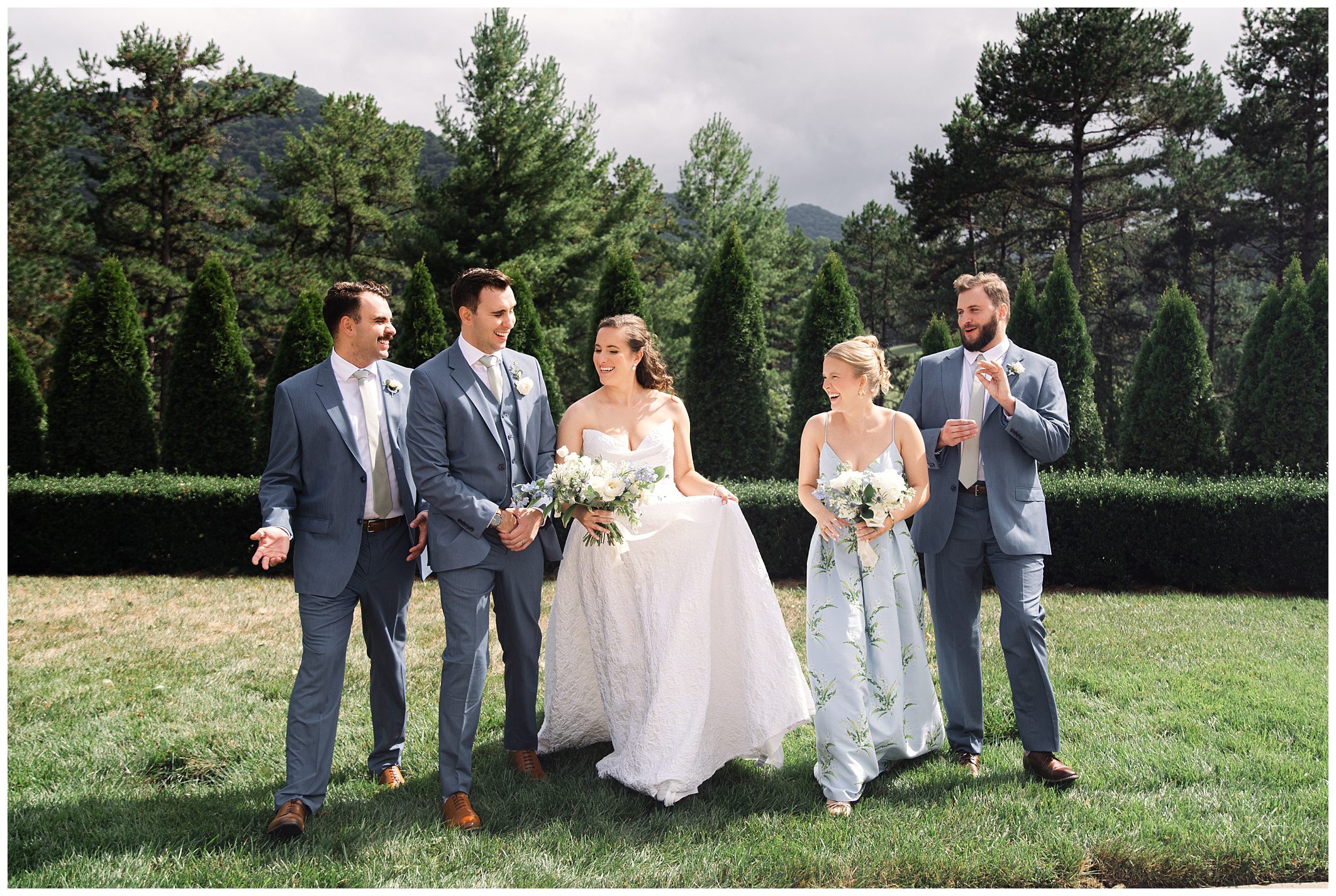 A bride and groom walk outdoors with three members of their wedding party, all wearing light blue attire. Trees and mountains are visible in the background.