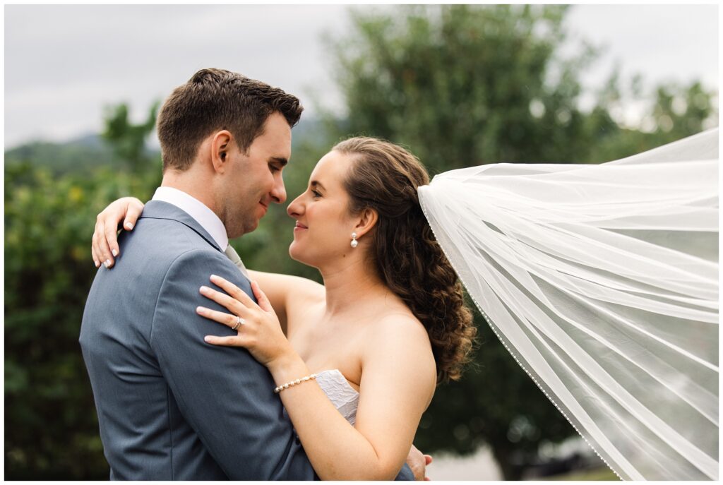 A bride and groom embrace outdoors; the groom wears a gray suit while the bride, in a strapless gown with a flowing veil, gazes into his eyes.