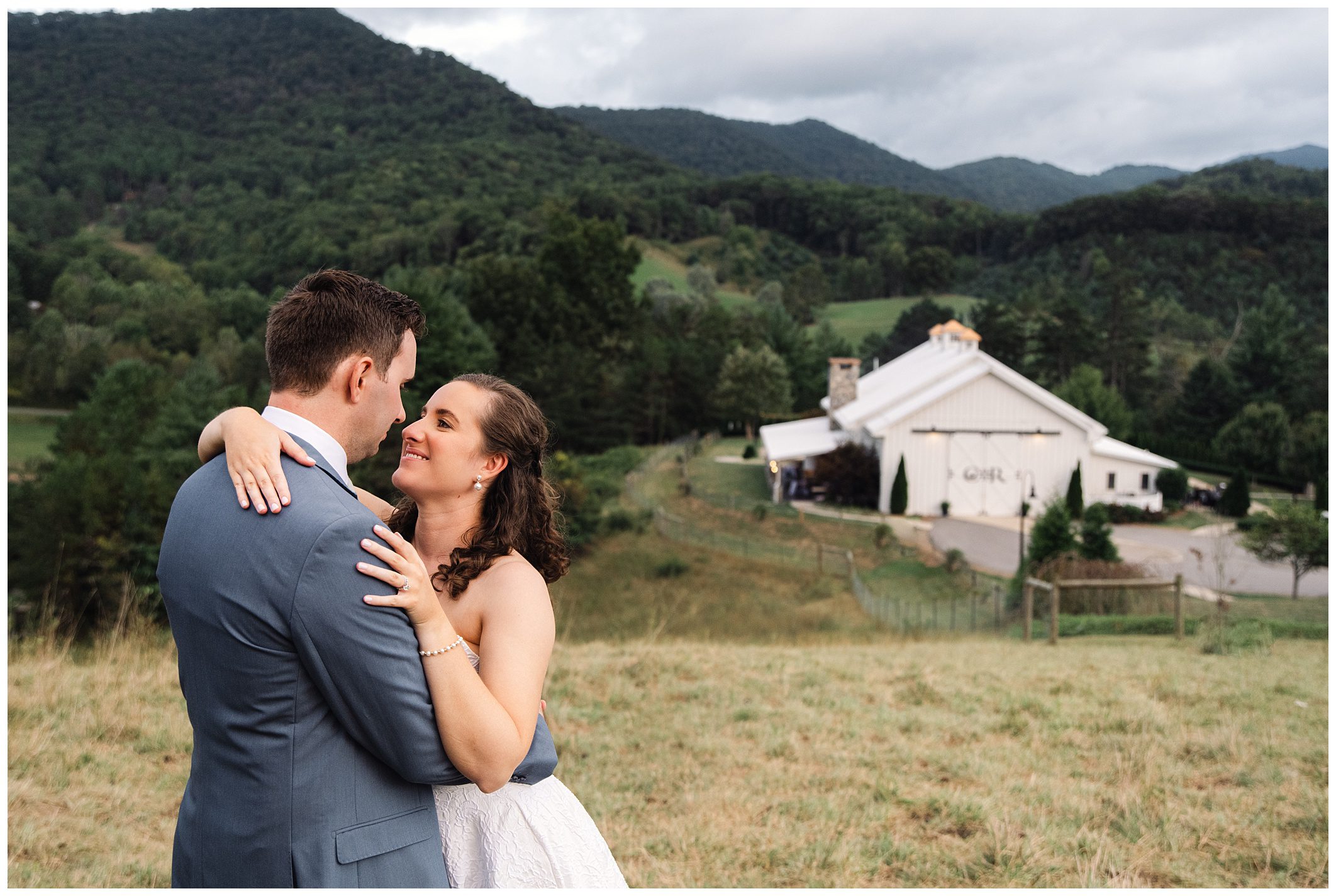 A couple in formal attire embraces on a grassy hill, with Chesnut Ridge in the Background at Mountain View wedding in blue.