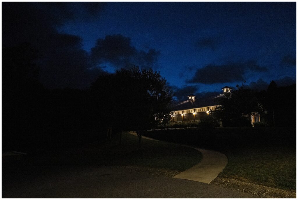 A well-lit building with two cupolas is visible against a dark night sky. A curved path leads up to the entrance, bordered by trees and a lawn.