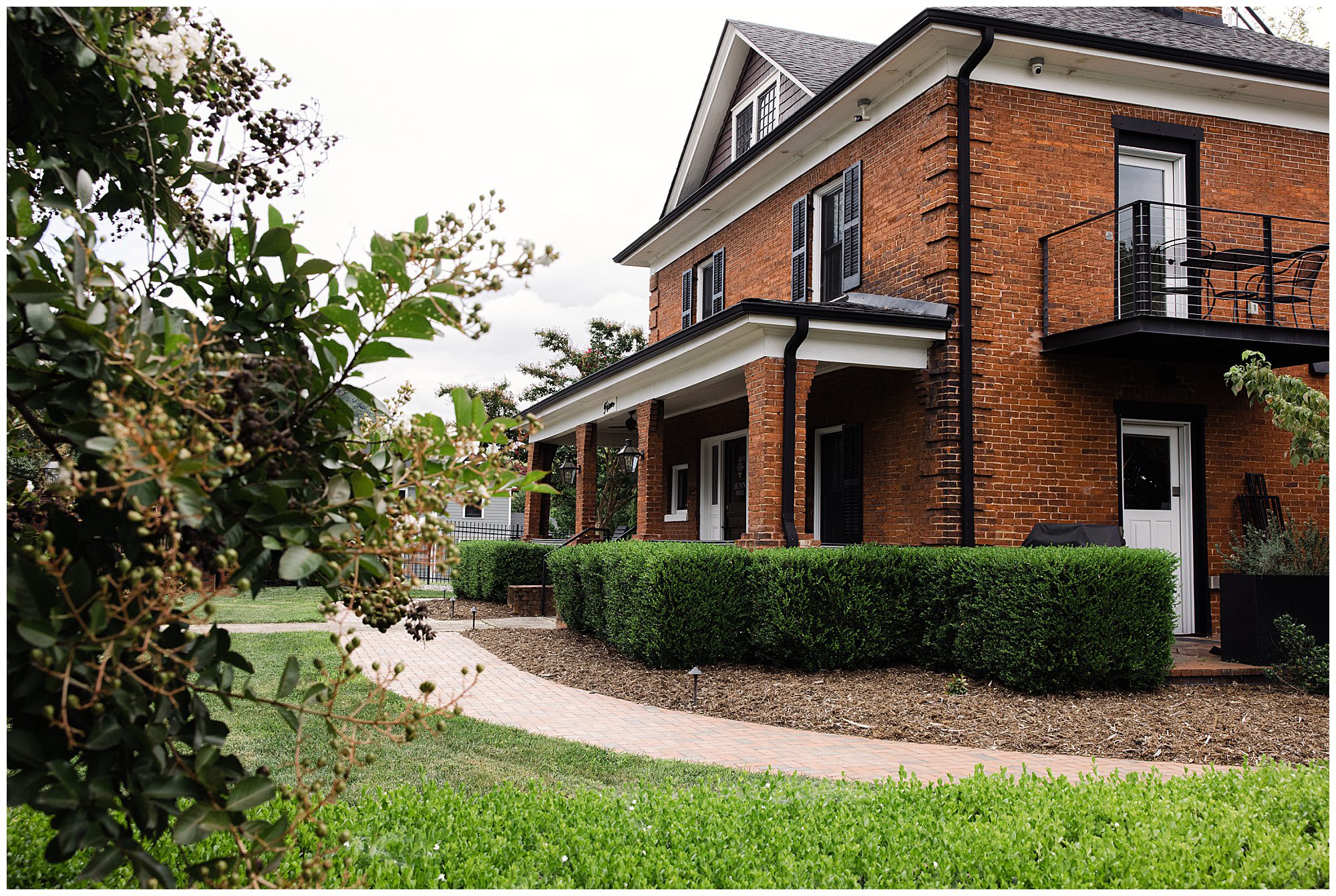 A two-story brick house with black shutters, a balcony, and a front porch, surrounded by green shrubs and a well-maintained lawn. A curved brick pathway leads to the front door.