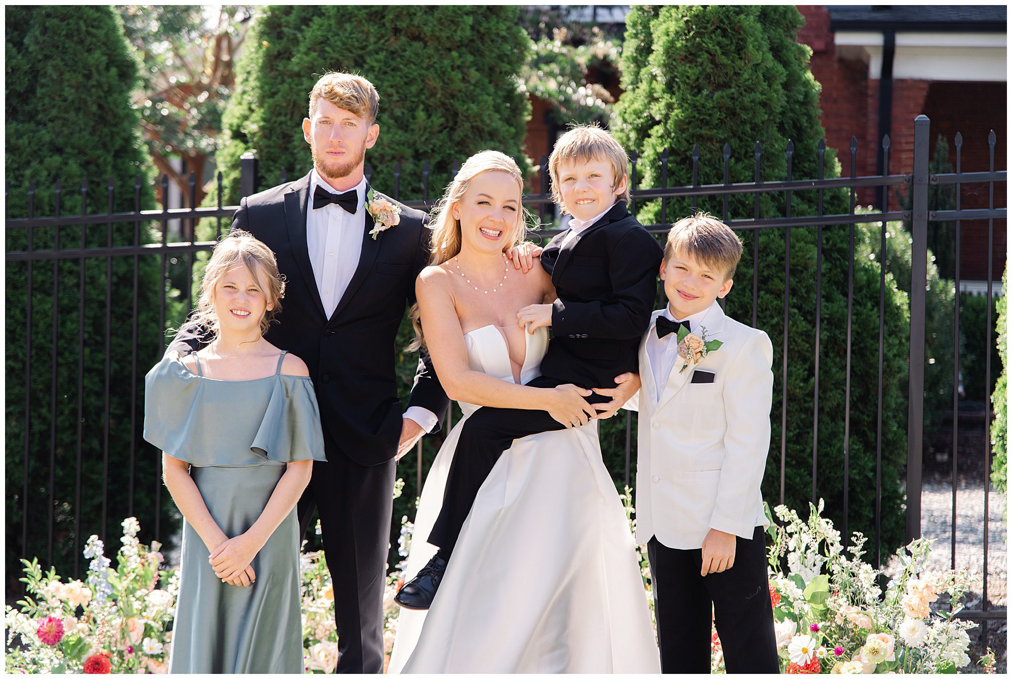 A newlywed couple stands smiling with three children, dressed formally, in front of a garden with tall greenery and a black iron fence. The bride is holding one of the children.