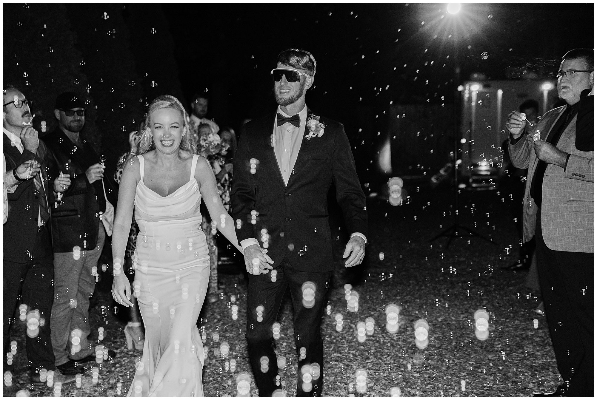 A bride and groom, holding hands, walk through a group of friends and family blowing bubbles during a nighttime celebration.