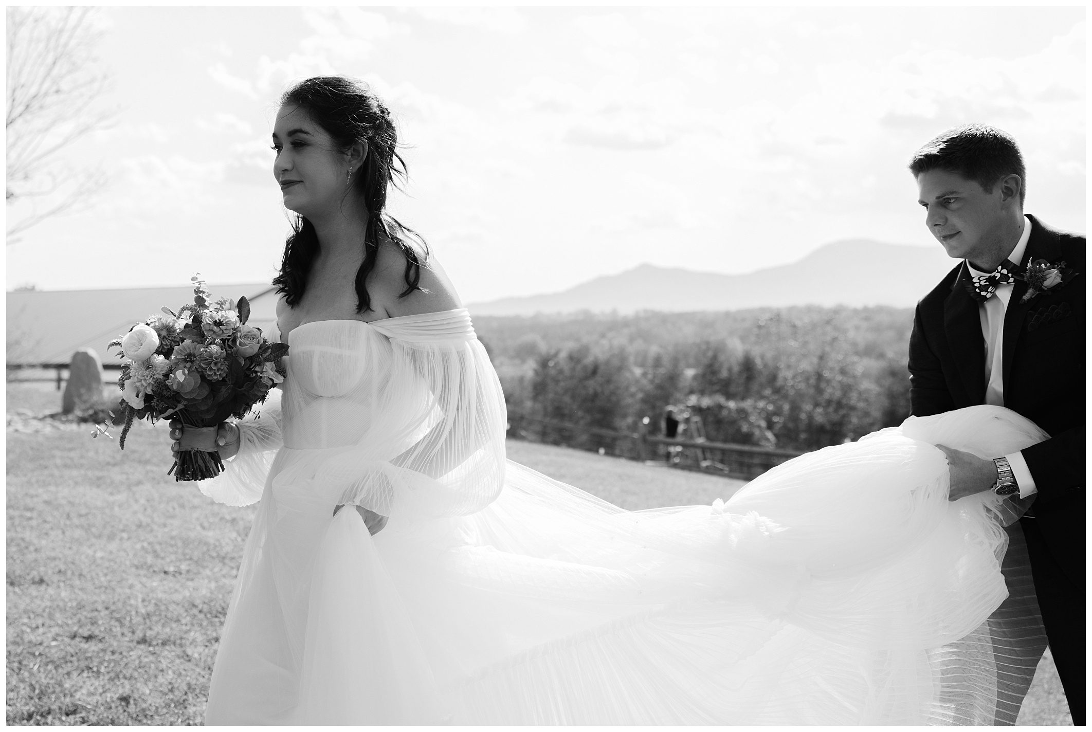 A bride in a flowing dress walks outdoors, holding a bouquet, while a person behind her carries her train.