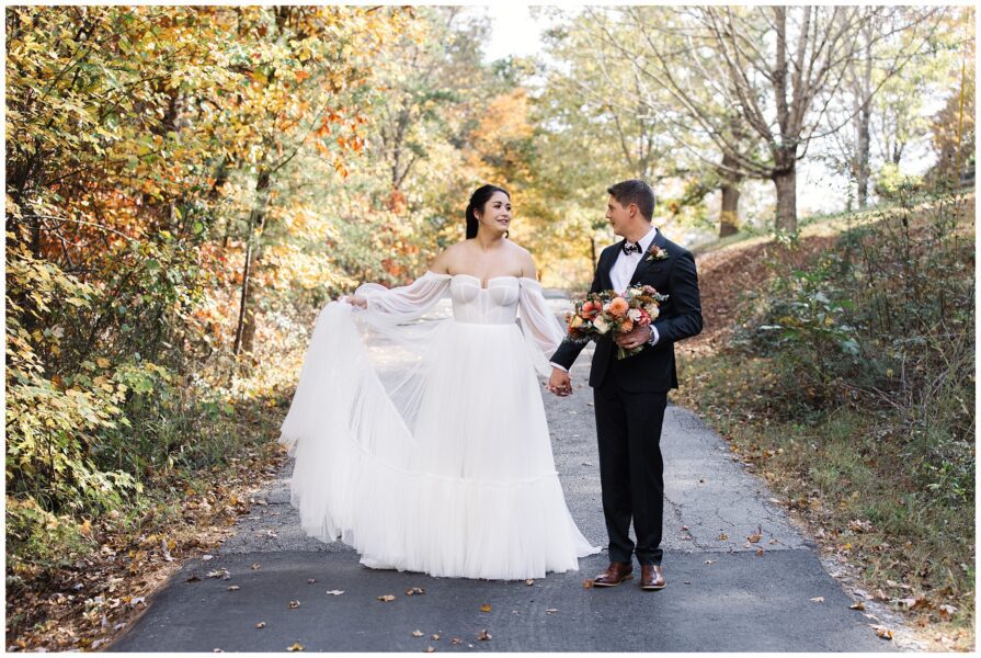 A bride and groom stand on a wooded path. The bride wears a white gown and holds part of the skirt; the groom wears a suit and holds a bouquet. The trees display fall foliage.