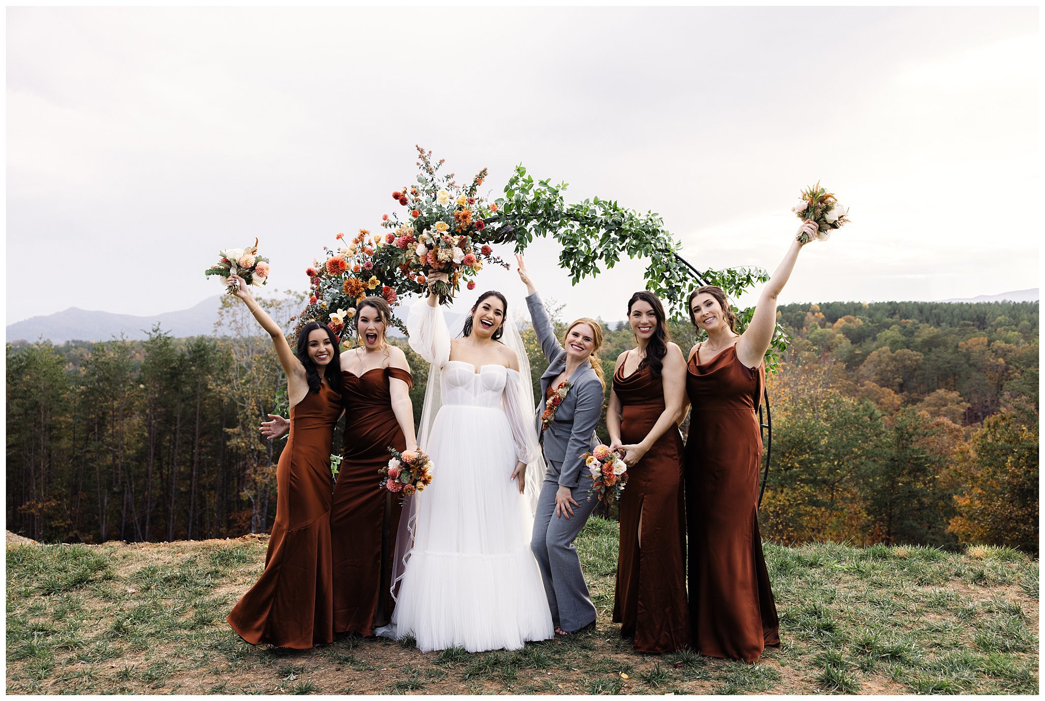 Bride and bridesmaids in brown dresses pose with bouquets in front of a floral arch outdoors.