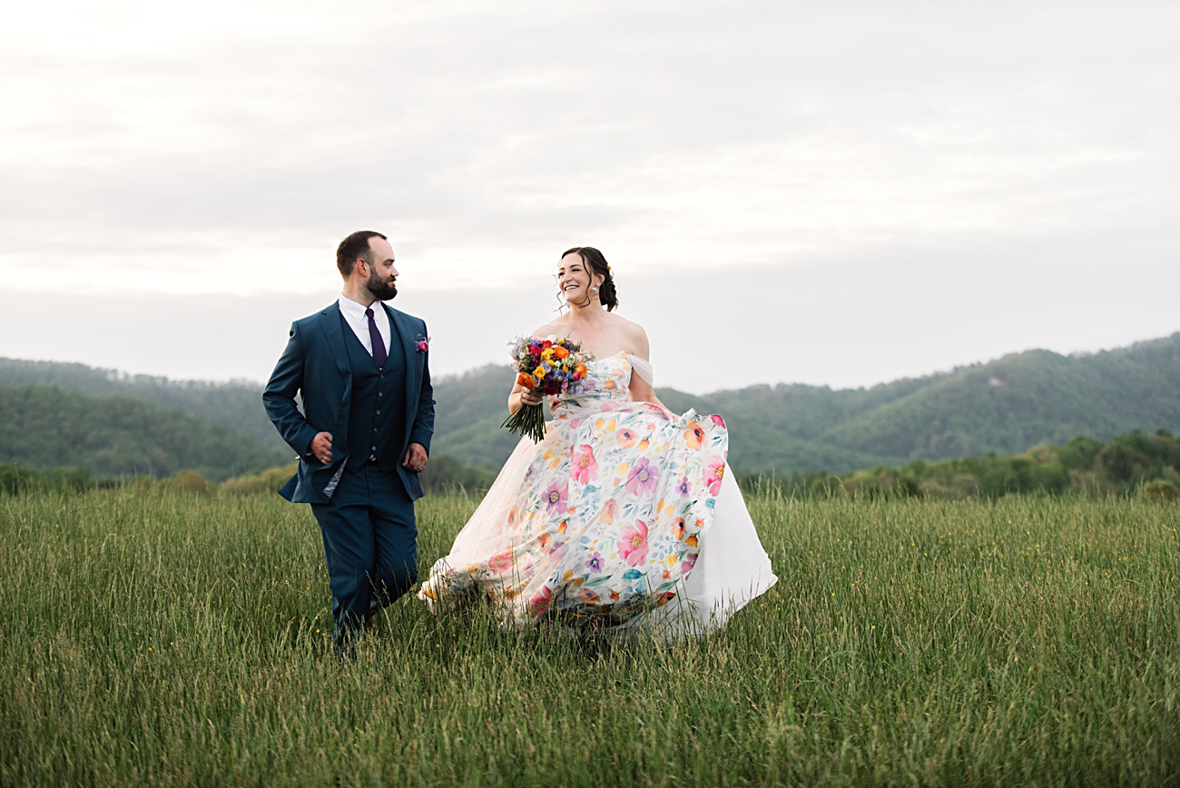 A bride in a colorful floral dress and a groom in a dark suit walk through a grassy field with hills in the background, capturing the charm of an Asheville NC Spring Garden Wedding.