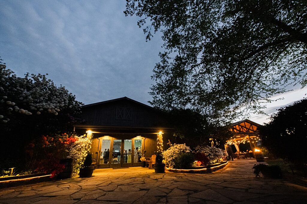 A warmly lit building and gazebo surrounded by trees and flowering bushes at dusk, with a stone pathway leading up to the entrance—perfect for a spring garden wedding in Asheville NC.