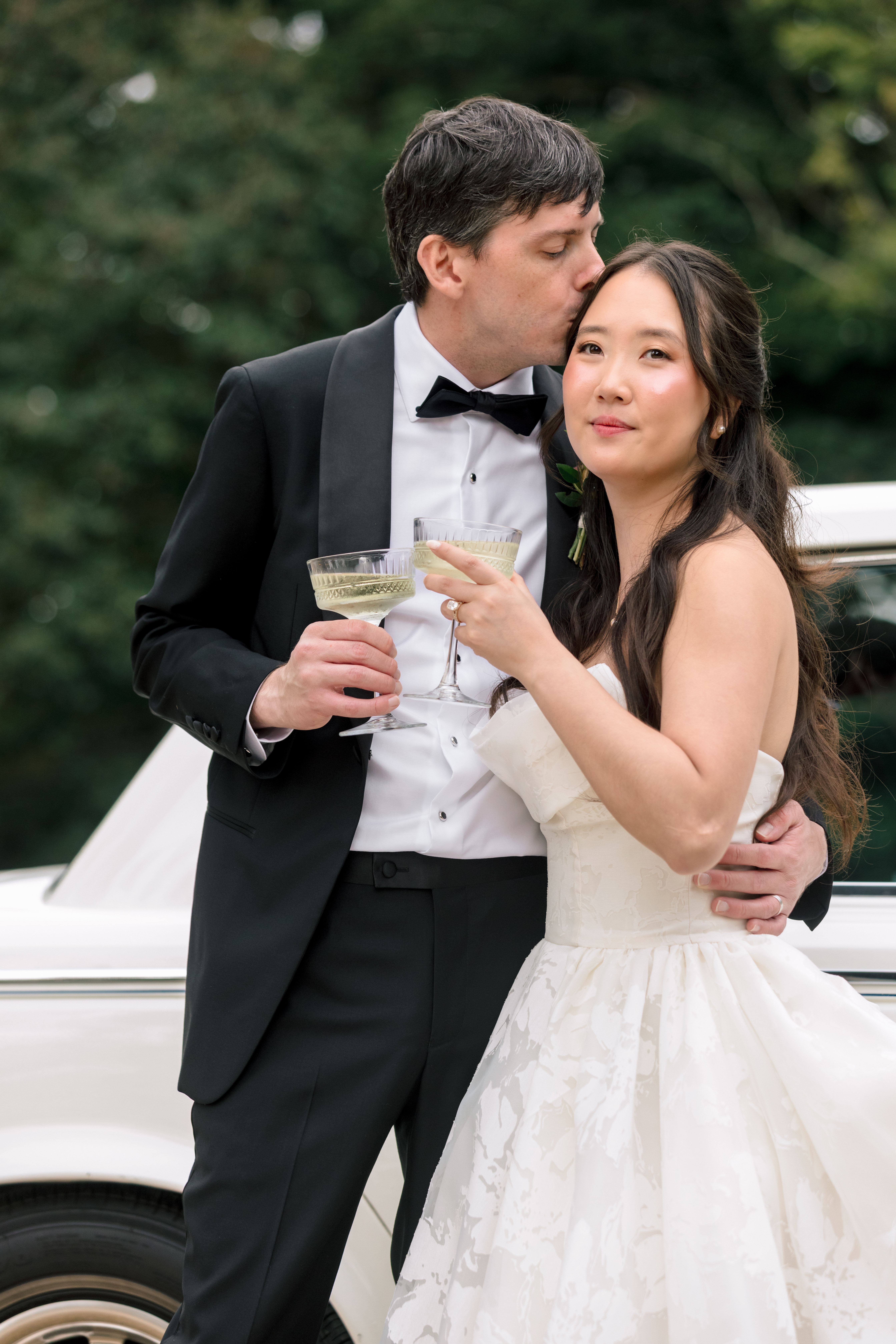 Bride and groom holding champagne coupes in front of a classic vintage getaway car at an Asheville North Carolina wedding celebration at The Farm.