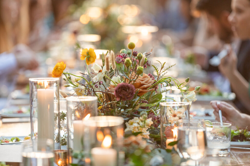 A close-up of a decorated dining table with flowers, candles, plates, and glasses, surrounded by people eating and conversing in soft, natural light—beautifully captured at High Holly wedding venue.