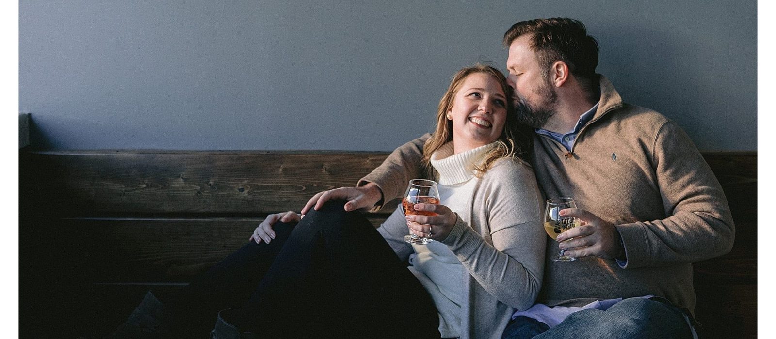 Young engaged couple sitting on wooden bench beside one another at urban orchard cidery in downtown Asheville, NC