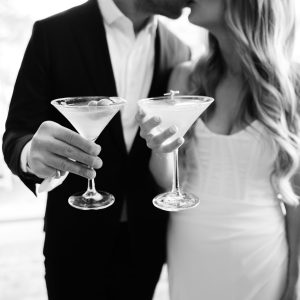 A couple dressed in formal attire kisses while each holds a martini glass; the black and white image captures the romantic moment, showcasing the artistry of an Asheville Wedding Photographer.