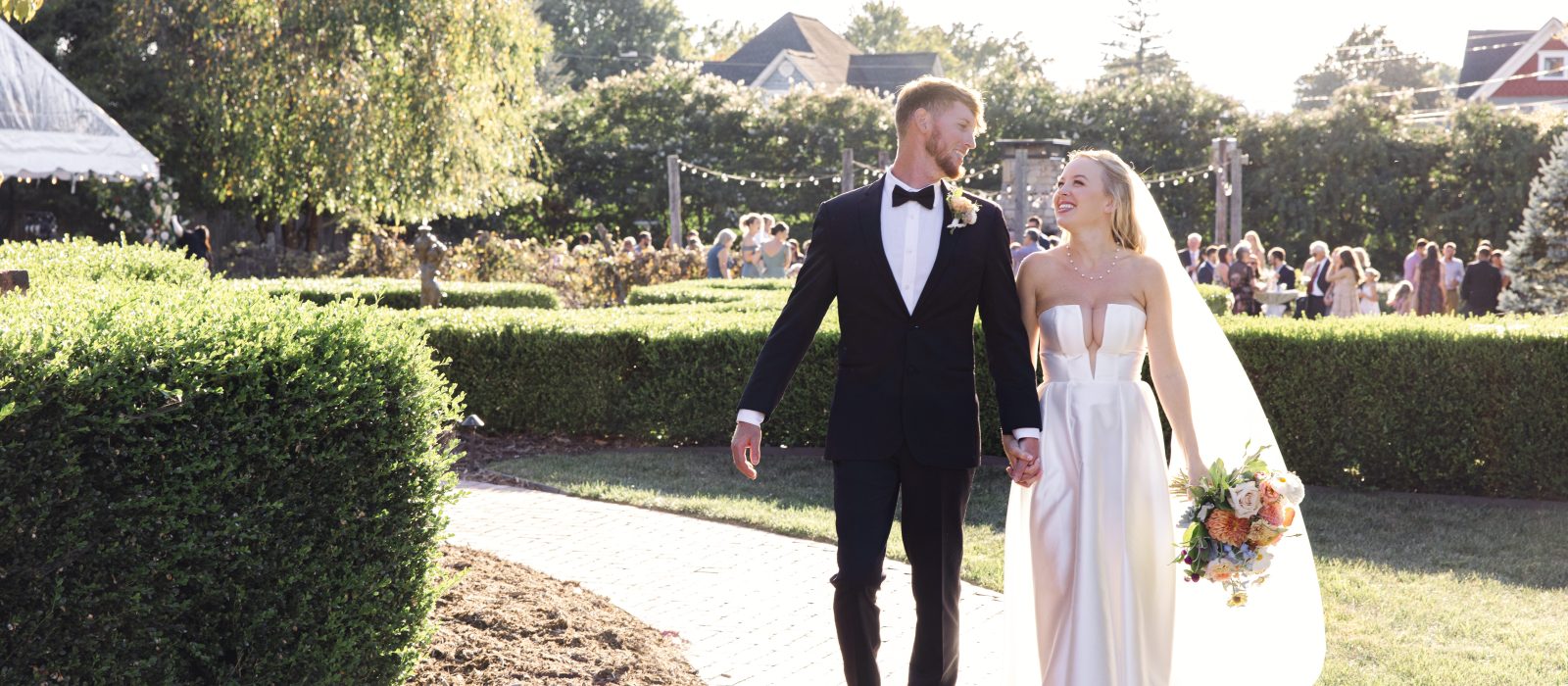 couple holding hands and walking together with guests at cocktail hour in the background