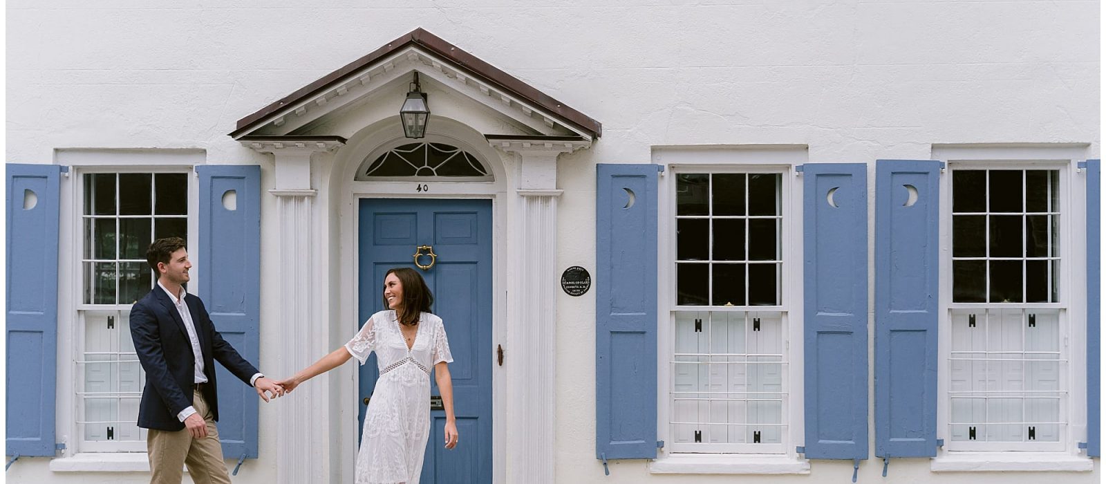young engaged couple walking holding hands infront of white home with blue shutters in downtown charleston