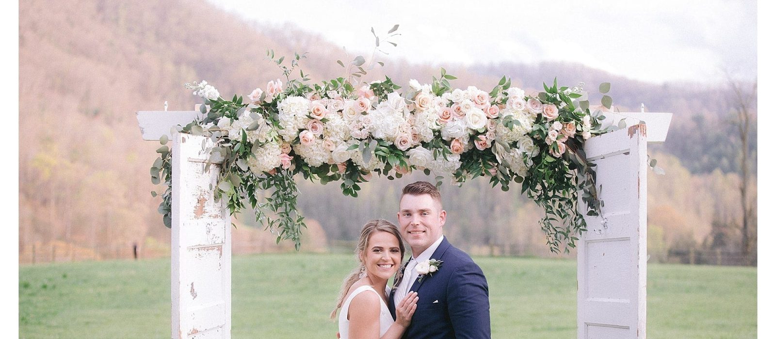 Bride and groom posing under wedding arbour of cream and peach flowers outdoors in grassy field