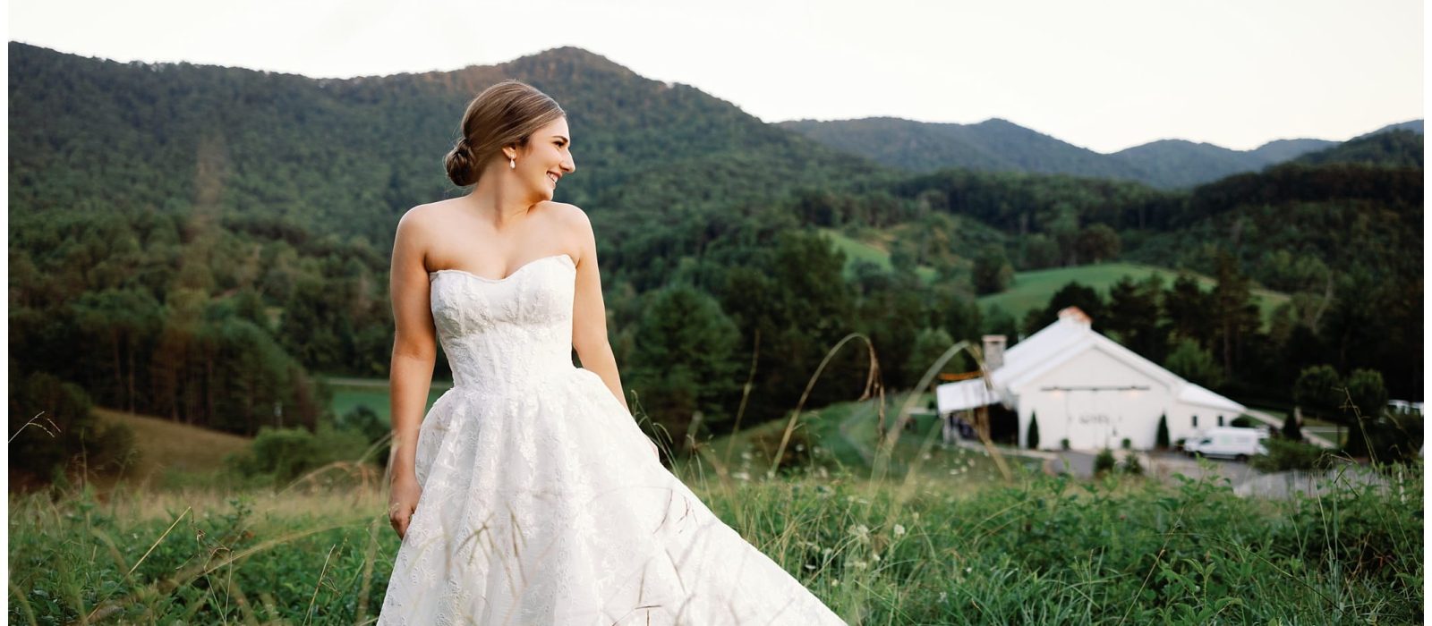 An elegant bride in a white wedding dress standing in a field with mountains as the backdrop for a summer wedding in Asheville.