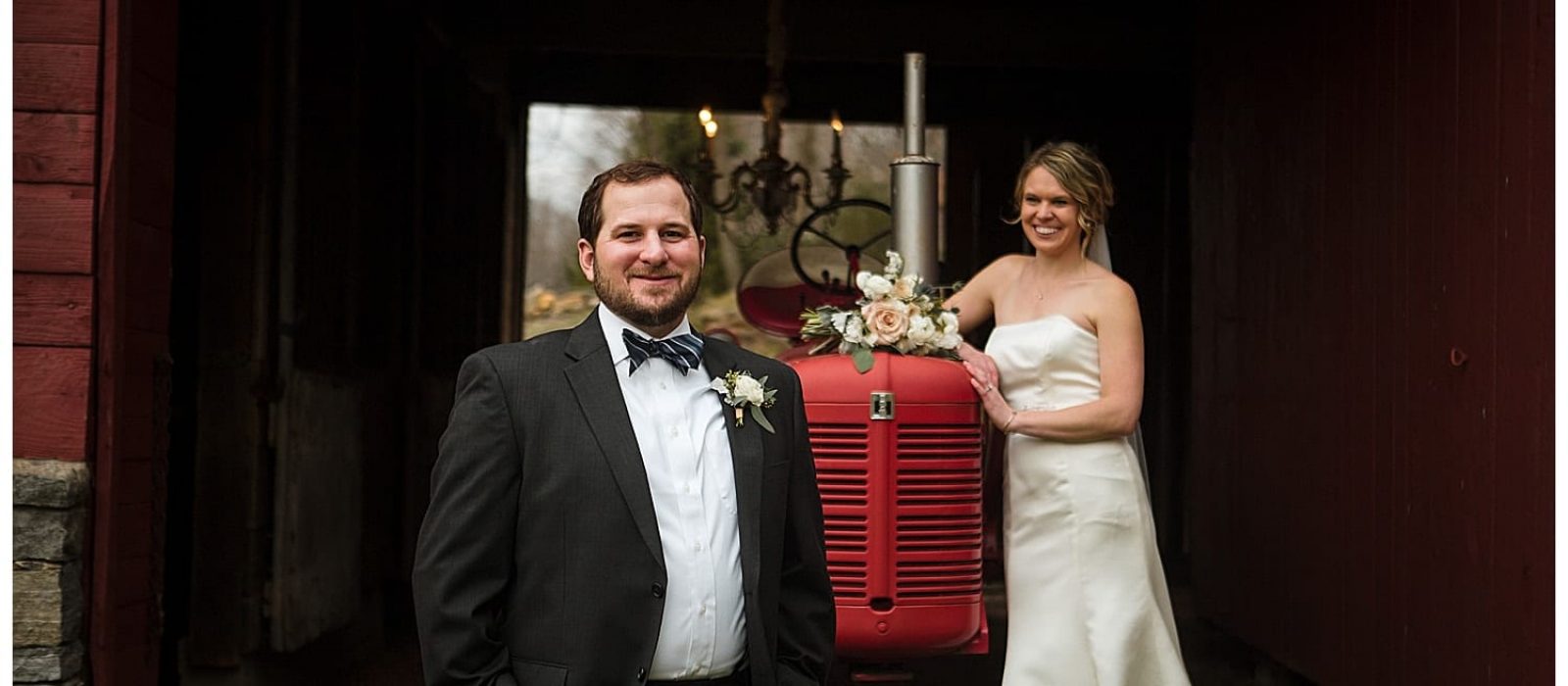 Bride and groom posing beside red rustic tractor, bride leaning against tractor smiling at groom while groom stands in front of tractor with hands in his pockets smiling and looking at camera