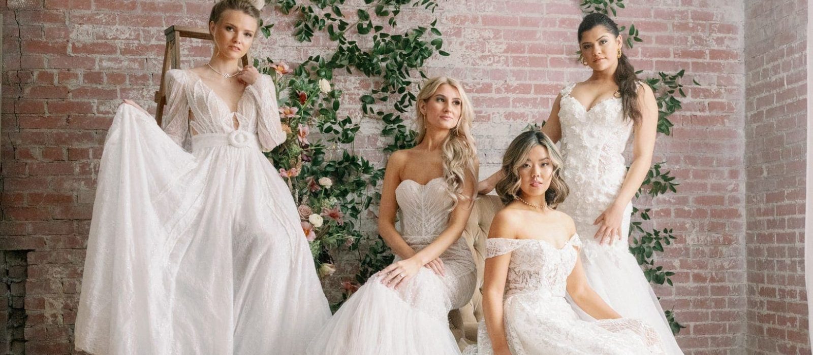 Four women wearing white vintage lace wedding gowns posing in front of light red brick wall with floral greenery running up it