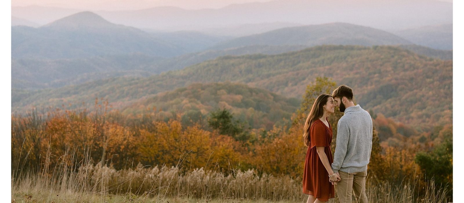 Young engaged couple kissing in golden grassy autumn field at sunset over looking mountain range
