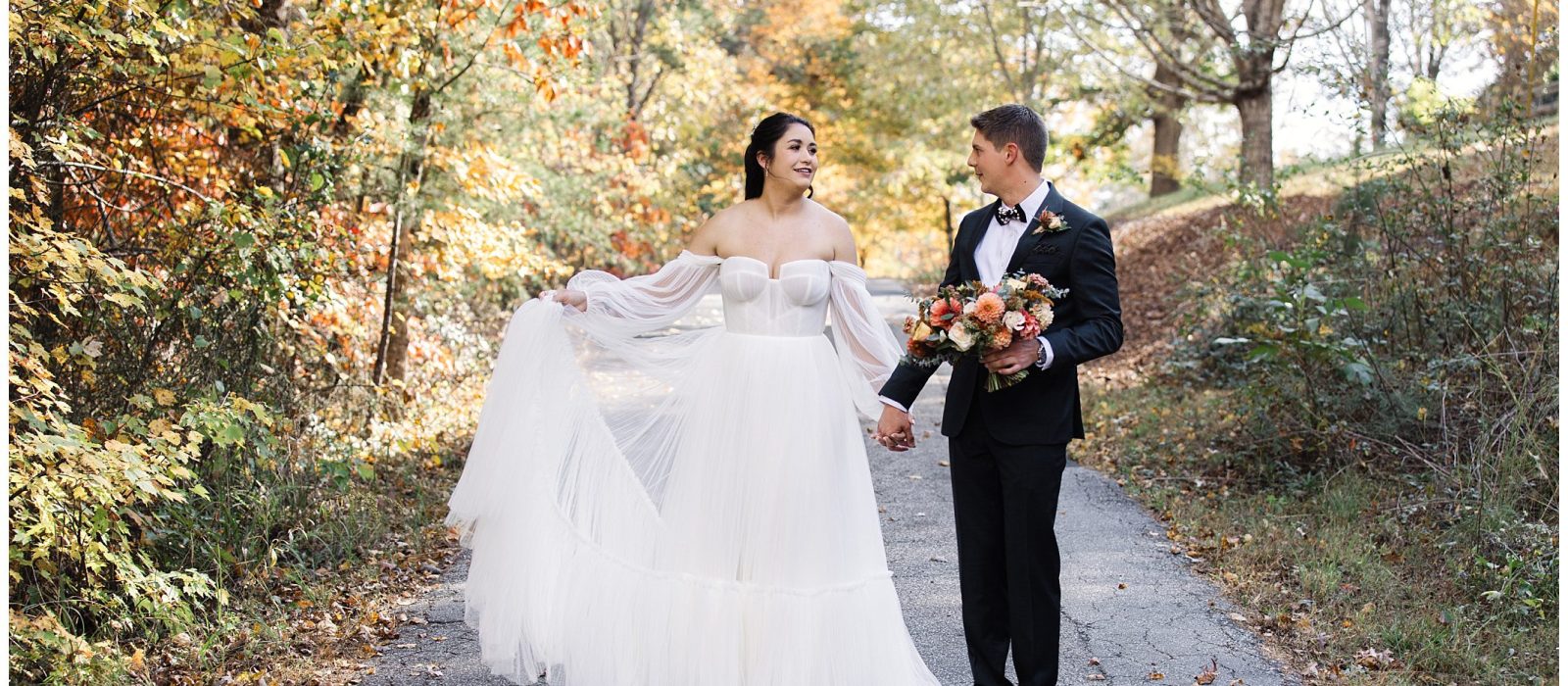 A bride and groom stand on a wooded path. The bride wears a white gown and holds part of the skirt; the groom wears a suit and holds a bouquet. The trees display fall foliage.