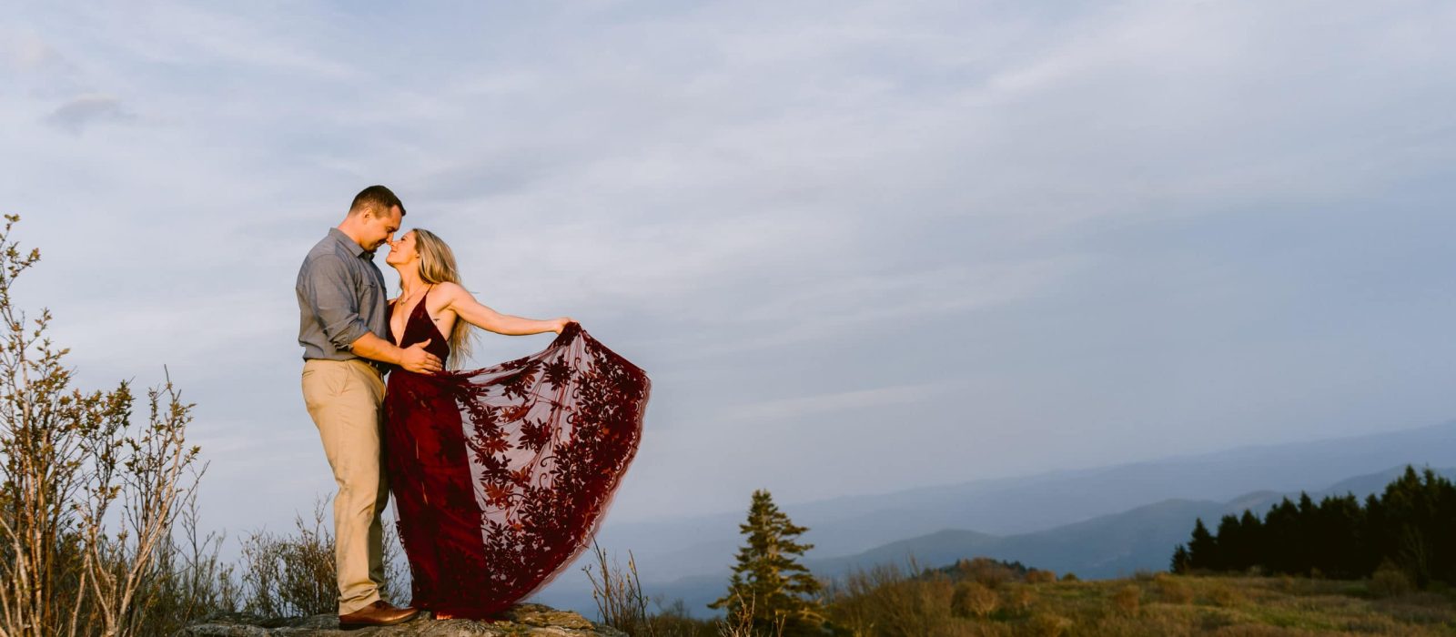 Engaged couple holding hands smiling at one another standing on rock surrounded by green trees and distant mountain views near Asheville, North Carolina photography done by Kathy Beaver.