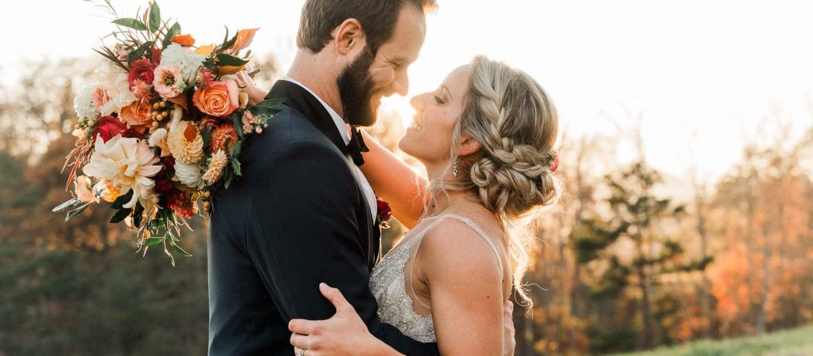 bride and groom standing on grassy field with sun setting behind them in the trees, arms around one another smiling about to kiss