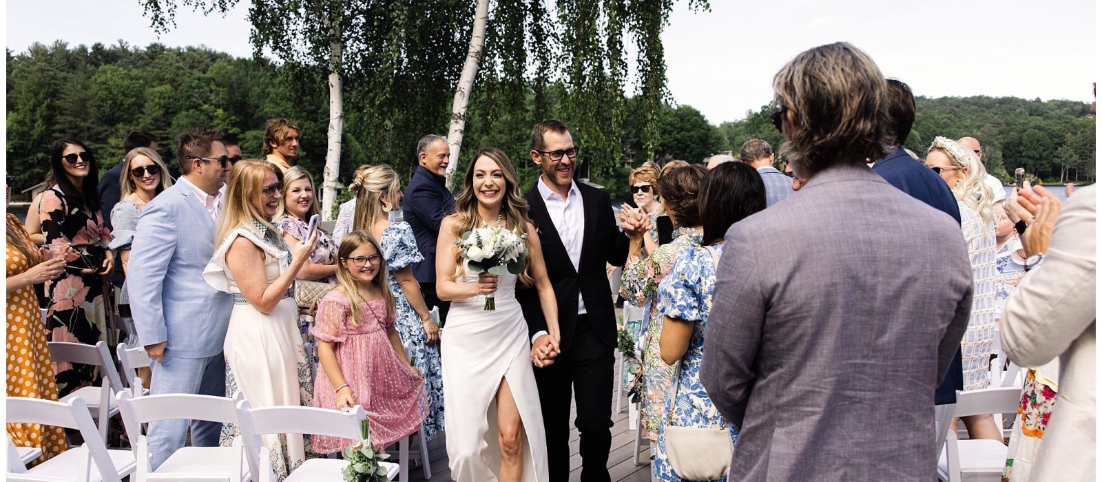 A bride and groom walk down an outdoor aisle surrounded by applauding guests. The bride holds a bouquet, and the groom wears a dark suit. There are trees in the background.