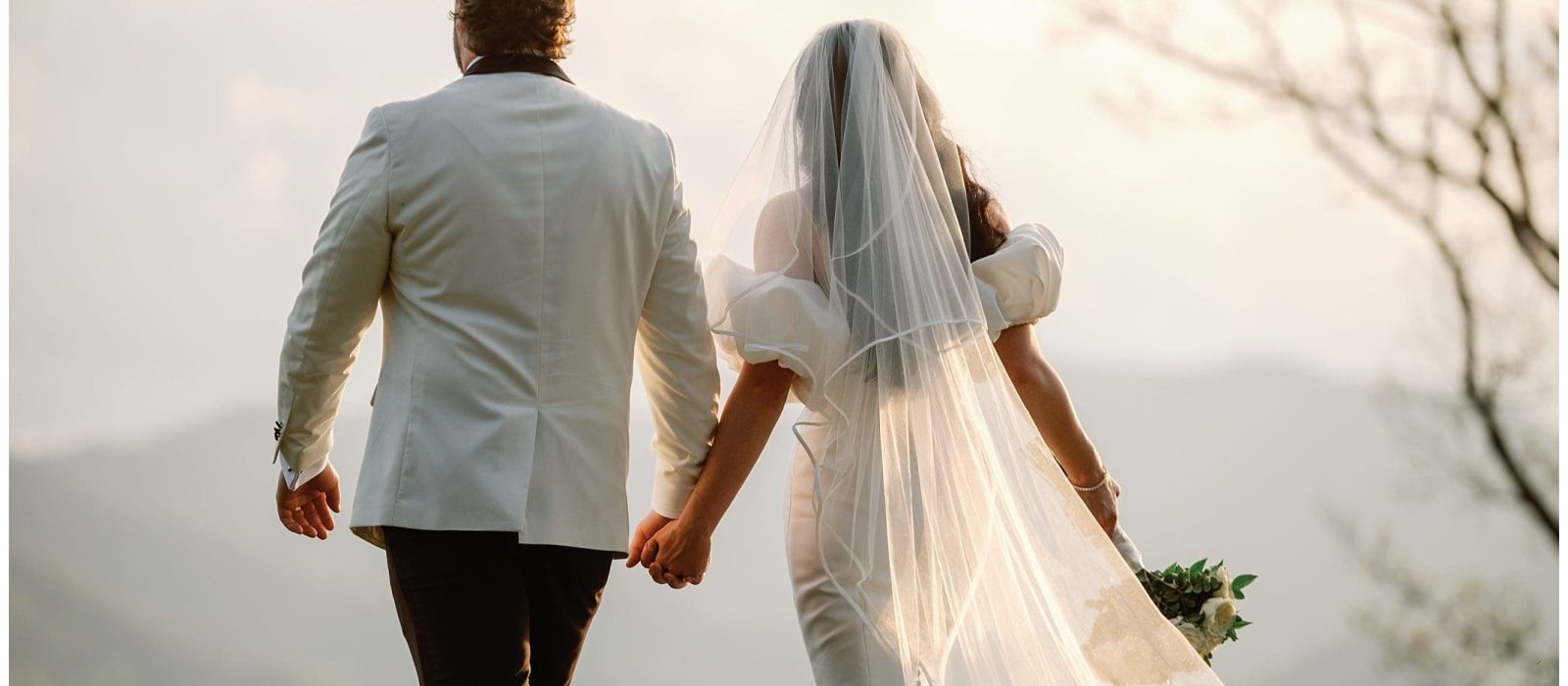 A bride and groom holding hands, walking toward a mountain vista, with the bride wearing a flowing veil and carrying a bouquet.
