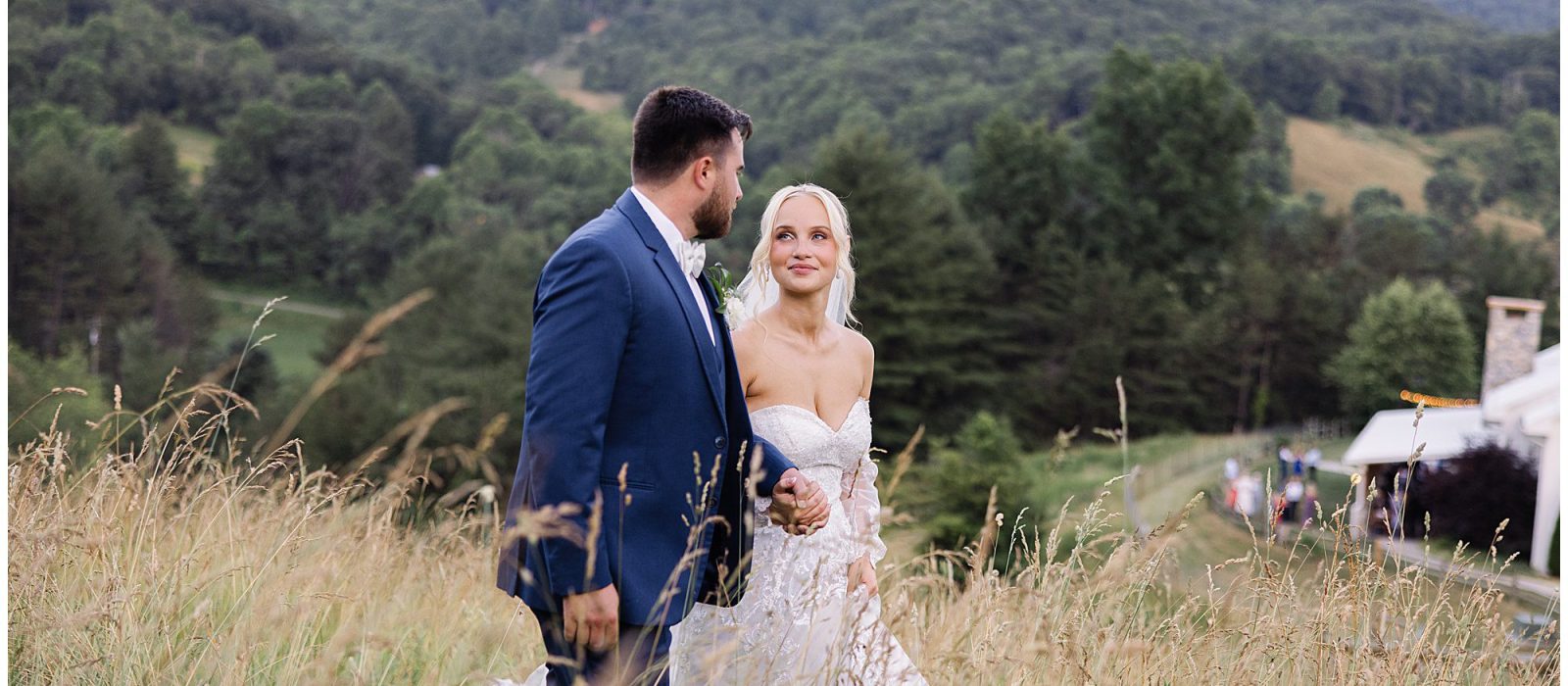 Bride and groom holding hands, walking through a grassy field with a forested mountain landscape in the background.