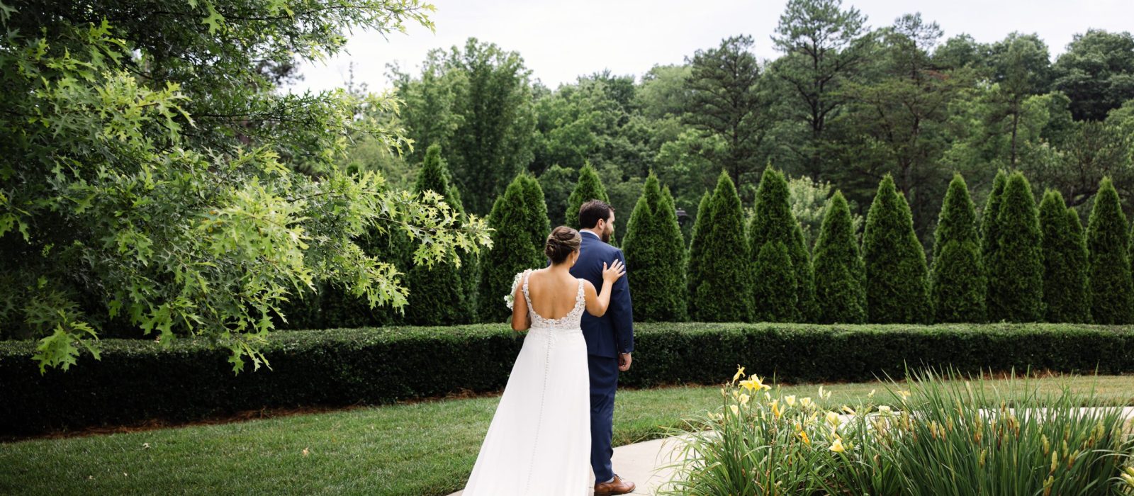 A bride and groom stand together on a path in a garden during their summer wedding in Asheville. The groom faces away while the bride touches his shoulder, surrounded by lush greenery and trees.