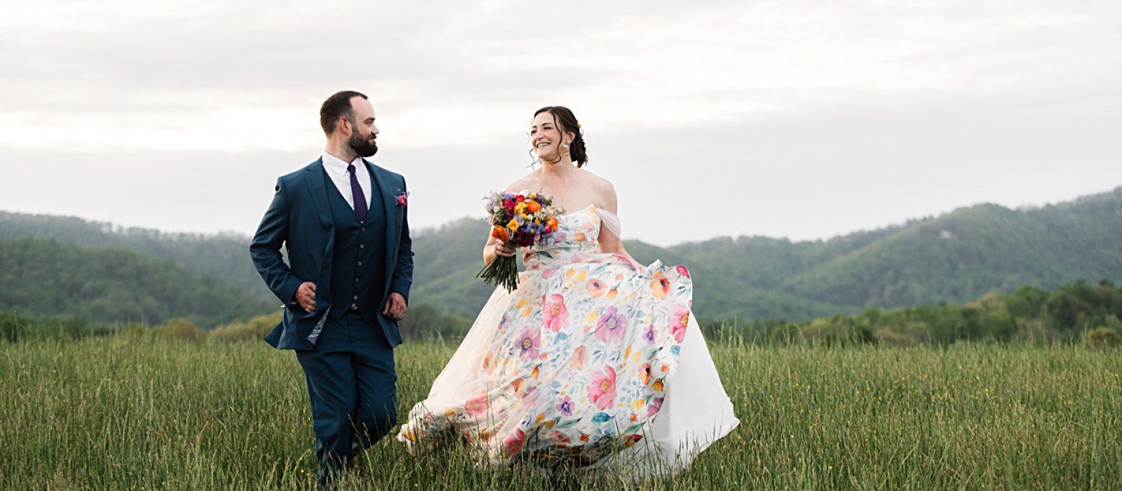 A bride in a colorful floral dress and a groom in a dark suit walk through a grassy field with hills in the background, capturing the charm of an Asheville NC Spring Garden Wedding.